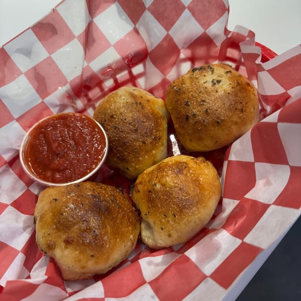 Four golden-brown garlic knots with herbs and a side of marinara sauce in a red checkered basket.